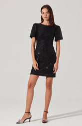 A woman is wearing a short black dress with a fitted silhouette. The dress features short, puffed sleeves and is covered in sparkly embellishments that add a shimmering effect. She pairs the dress with black high-heeled sandals that have ankle straps and decorative bows on the front.